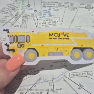 May include: A yellow fire truck sticker with the words "Mojave Air and Space Port" printed on the side. The sticker is on a map with a compass rose and other airport markings.