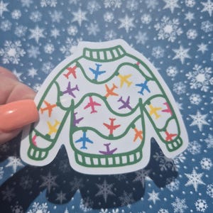 Puede incluir: Una pegatina blanca con forma de jersey, decorada con aviones de colores. El jersey tiene un contorno verde y un cuello y puños acanalados. Los aviones son de varios colores, incluyendo rojo, naranja, amarillo, morado y azul.