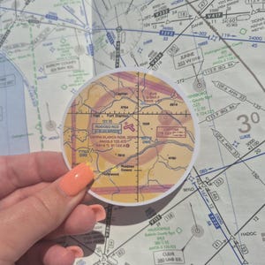May include: A circular sticker with a map of the area around Ruidoso, New Mexico. The sticker is yellow with red and pink accents. The map shows the location of Ruidoso, Capitan, Fort Stanton, and Hollywood. The sticker is designed for pilots and includes the text "RUIDOSO ACO ALBUQUERQUE" and "SERRA BLANCA RGNL ISPRU AWOS-3 126.475 3414.5 8122.8".