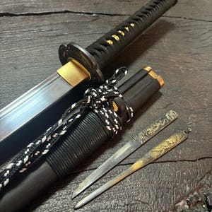 Puede incluir: Una espada katana japonesa con empuñadura y vaina negras, acentuada con detalles dorados. La hoja es oscura, y también hay dos pequeños cuchillos decorativos con diseños ornamentados.