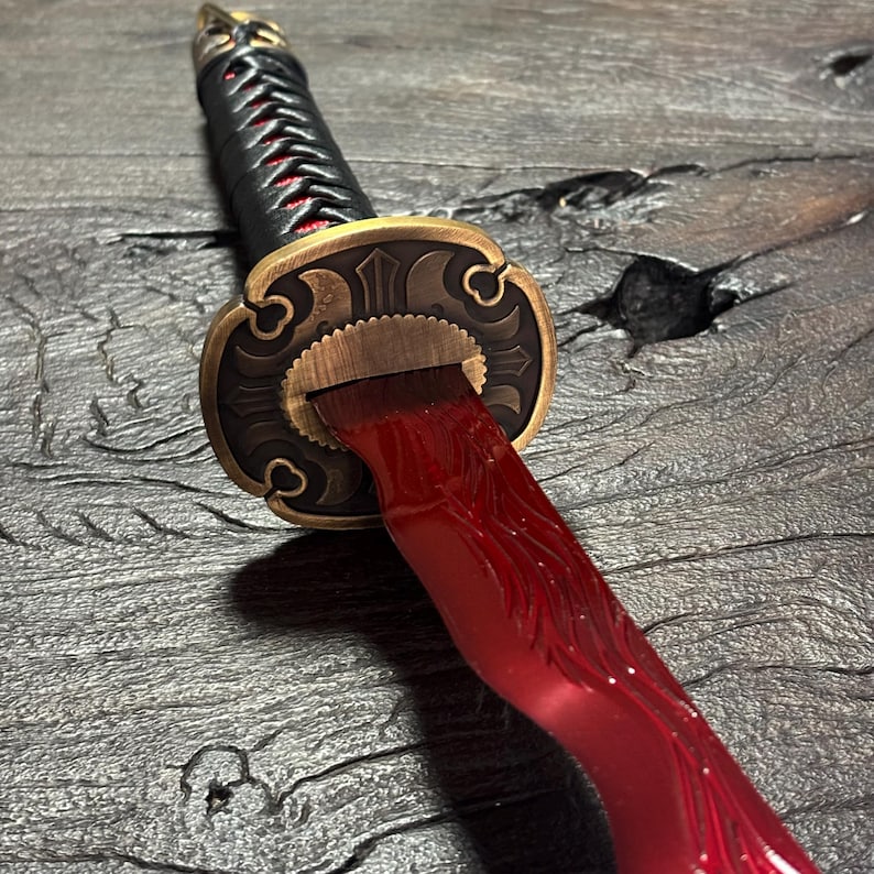 May include: Close-up of a katana sword with a red blade and a black handle wrapped with red cord. The guard is bronze-colored with intricate designs. The sword rests on a dark, textured surface.