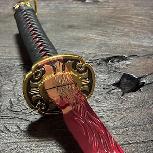 May include: A detailed view of a katana sword. The handle is wrapped in black material with red accents. The guard is gold with intricate designs, and the blade is a vibrant red color.