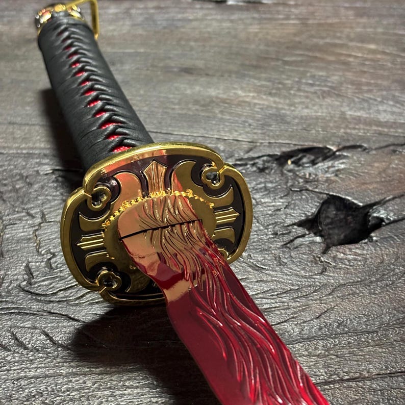 May include: A detailed view of a katana sword. The handle is wrapped in black material with red accents. The guard is gold with intricate designs, and the blade is a vibrant red color.