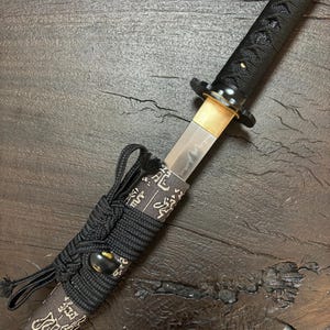 Op de afbeelding: Close-up van een Japanse tanto-mes. Het lemmet is zilverkleurig, met een goudkleurige greep en een zwarte handgreep gewikkeld in een textuurpatroon. De schede is zwart met goudkleurige Japanse karakters. Het is een traditioneel Japans wapen.