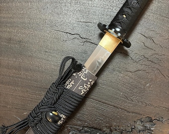May include: Close-up of a Japanese tanto knife. The blade is silver, with a gold-colored hilt and a black handle wrapped in a textured pattern. The scabbard is black with gold-colored Japanese characters. The knife is a traditional Japanese weapon.