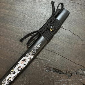 May include: A black katana scabbard with a detailed white dragon design. The scabbard is wrapped with black cord and features a decorative knot. The design is a traditional Japanese art style.