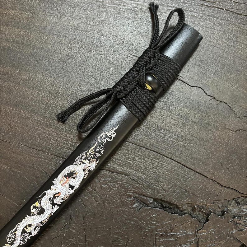 May include: A black katana scabbard with a detailed white dragon design. The scabbard is wrapped with black cord and features a decorative knot. The design is a traditional Japanese art style.