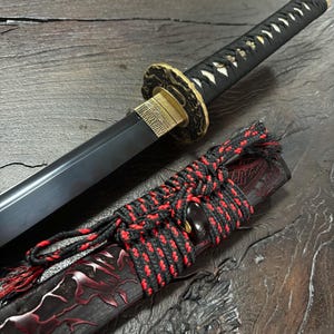 May include: A black katana sword with a gold-colored guard and a black handle wrapped in white and black fabric. The scabbard is black with red accents and a red and black cord.