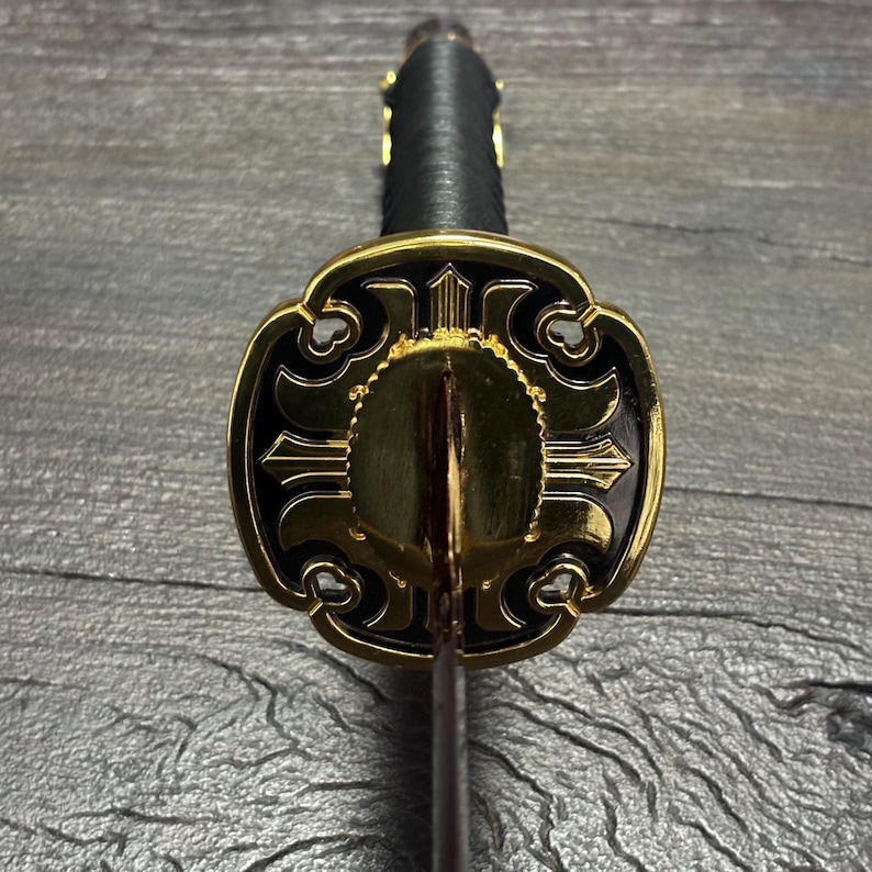 May include: Close-up of a katana sword. The guard is gold and black with an ornate design. The handle is wrapped in black material, and the blade is visible. The sword is set against a dark wood grain background.
