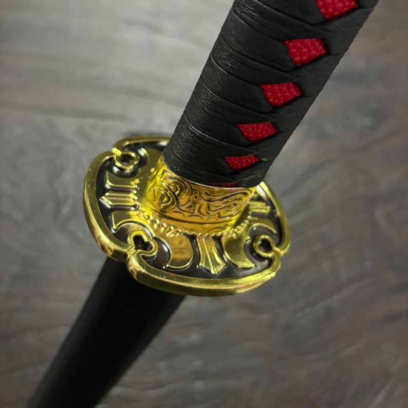 May include: Close-up of a katana sword hilt. The handle is wrapped in black fabric with red accents. A gold-colored guard with intricate designs separates the handle from the black blade.