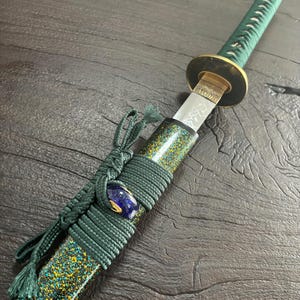 May include: A detailed view of a Japanese katana sword. The handle and scabbard are wrapped in green cord. The scabbard has a speckled pattern of gold and teal. A blue and gold bead accents the handle. The blade is visible.