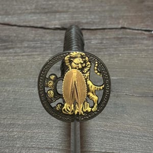 May include: Close-up of a decorative sword hilt. The hilt features a circular guard with a detailed lion and foliage design in gold and black. The handle is wrapped in a dark, textured material.