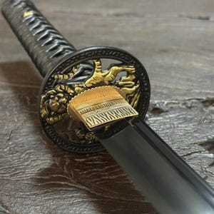 May include: Close-up of a Japanese katana sword. The handle is wrapped in a dark gray material, and the guard features gold-colored dragon detailing. The blade is black and reflective. The sword is a traditional weapon.
