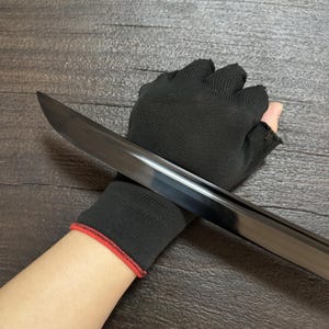 May include: A black sword blade with a polished finish. The sword is held by a black glove with a red trim. The blade is angled across the image, reflecting light. The background is a dark wood grain.