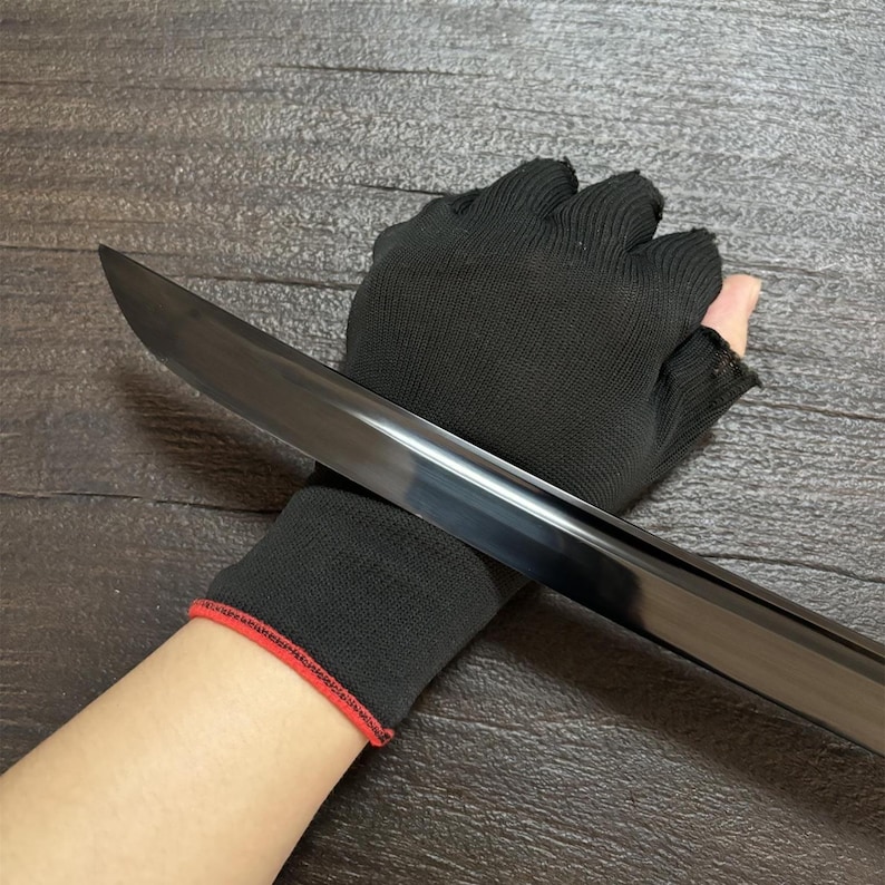 May include: A black sword blade with a polished finish. The sword is held by a black glove with a red trim. The blade is angled across the image, reflecting light. The background is a dark wood grain.