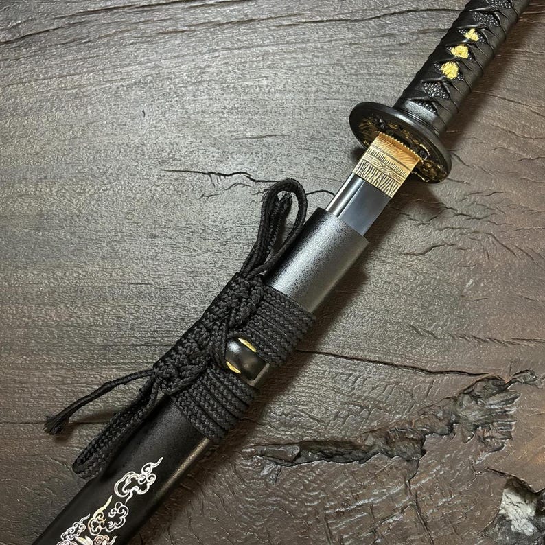 May include: A black katana with gold accents. The handle is wrapped in black fabric, and the blade is partially visible. The scabbard is black with ornate silver designs. The sword is displayed on a dark wooden surface.