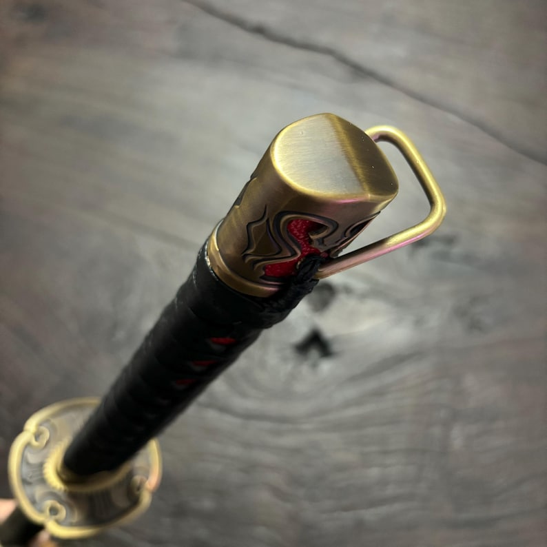 May include: Close-up of a katana sword. The handle is wrapped in black material, with a gold-colored metal guard and accents. The end cap features a decorative design with red accents. The sword's blade is not visible.