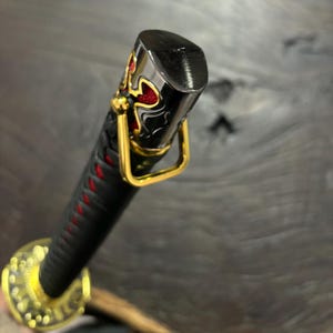 May include: Close-up of a katana handle. The black handle is wrapped with red stitching. The pommel is black with gold and red decorative accents. A gold-colored metal ring is attached to the handle.
