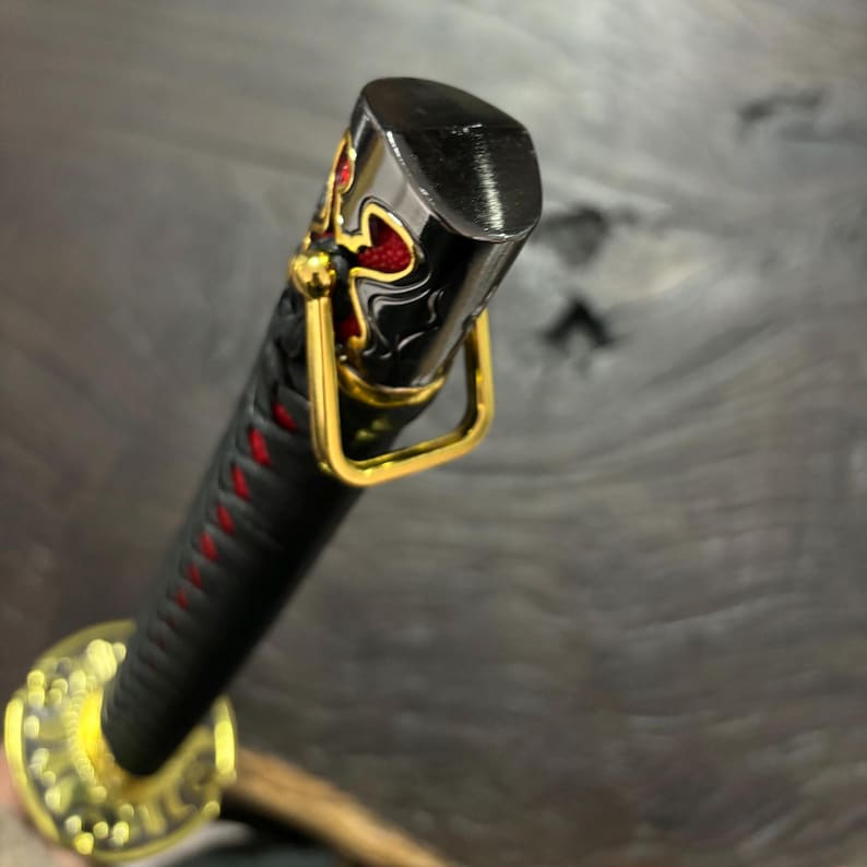 May include: Close-up of a katana handle. The black handle is wrapped with red stitching. The pommel is black with gold and red decorative accents. A gold-colored metal ring is attached to the handle.