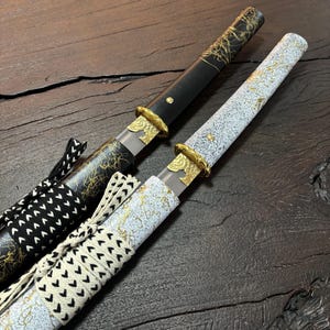May include: Two Japanese-style swords with sheaths. One sheath is black with gold detailing, the other is white with gold speckles. Both have gold-coloured fittings and decorative cord wrapping. The blades are visible within the sheaths.