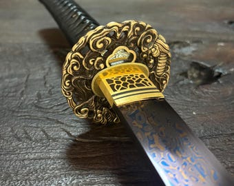 May include: Close-up of a Japanese katana sword. The blade has a dark finish with blue and gold patterns. The guard is ornate with gold and bronze detailing. The handle is black and textured.