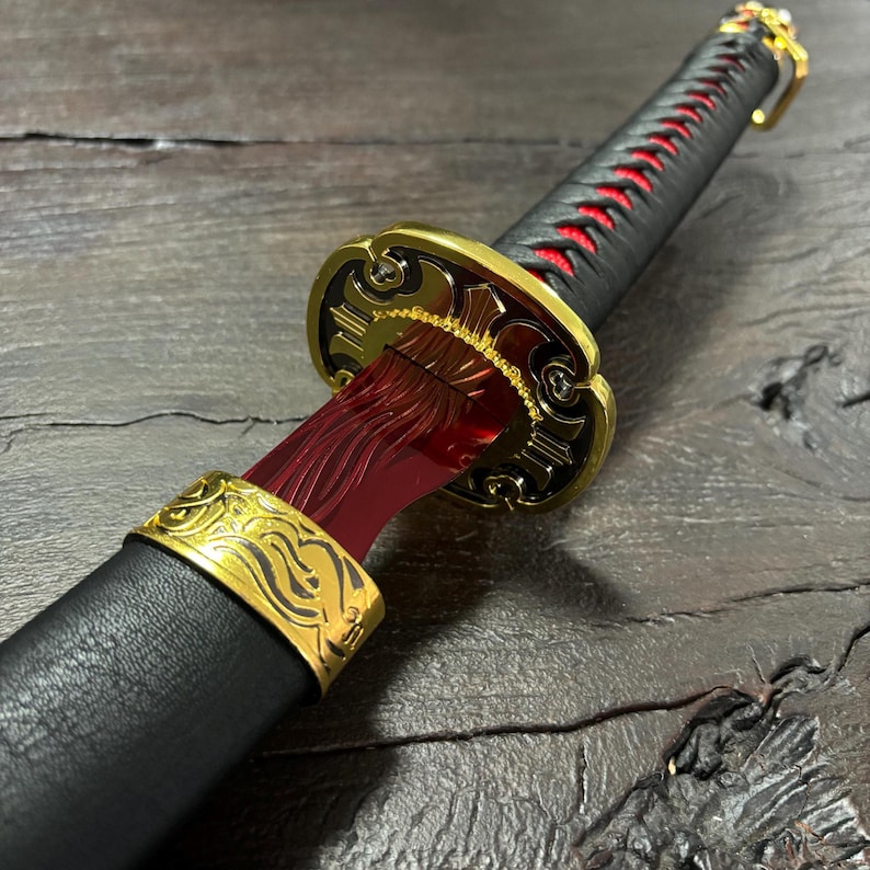 May include: A detailed close-up of a decorative sword. The sword features a black handle with red stitching, a gold-colored guard, and a red blade. The blade is visible through the guard. The sword is on a dark wooden surface.