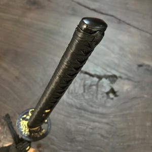 May include: Close-up of a katana handle. The handle is wrapped in black material with a textured pattern. The guard features gold-colored accents. The pommel is black and rounded.