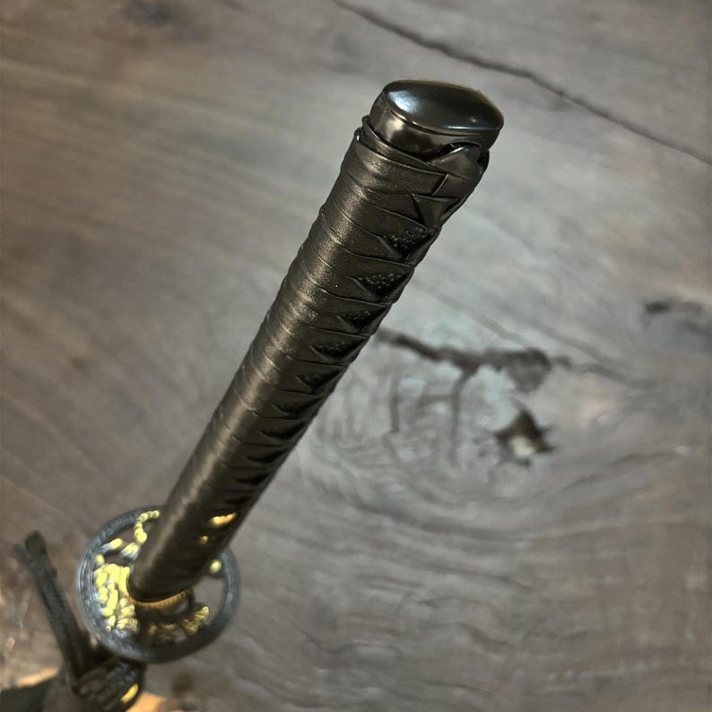 May include: Close-up of a katana handle. The handle is wrapped in black material with a textured pattern. The guard features gold-colored accents. The pommel is black and rounded.