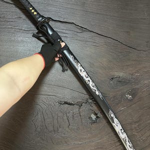 May include: A black katana sword with a decorative silver dragon design along the blade. The handle is wrapped in black fabric, and the scabbard is black. The sword is displayed on a dark wooden surface.