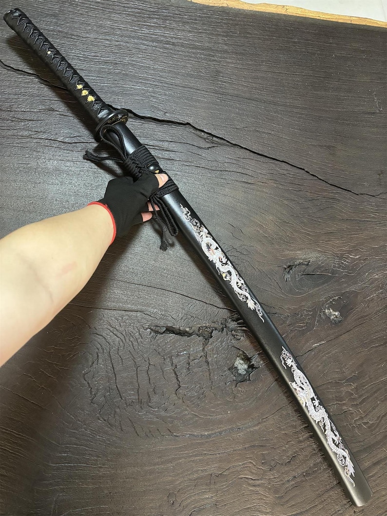 May include: A black katana sword with a decorative silver dragon design along the blade. The handle is wrapped in black fabric, and the scabbard is black. The sword is displayed on a dark wooden surface.