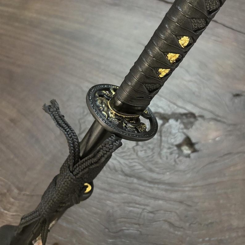 May include: Close-up of a katana sword. The handle is wrapped in black leather with gold accents. The guard is black with gold detailing. A black cord is attached to the handle. The blade is not visible.