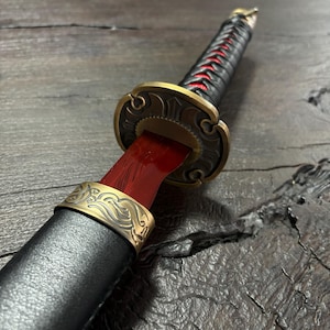 May include: Close-up of a decorative katana sword. The sword features a black handle wrapped with red cord, a brass-colored guard, and a red blade. The scabbard is black with a brass-colored band.