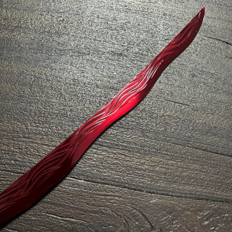 May include: A long, curved, red object with a pointed tip. The surface is carved with a wave-like pattern. The object is set against a dark, textured background.