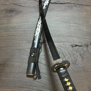 May include: A black katana sword with a decorative handle and scabbard. The scabbard features a white dragon design. The handle is wrapped in black material with gold accents. The blade is black.