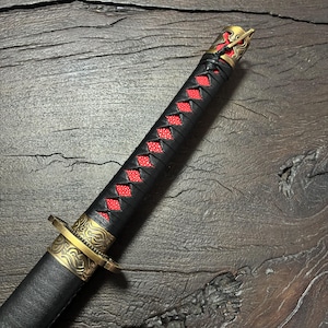 May include: Close-up of a katana sword. The handle is wrapped in black leather with red diamond-shaped accents. The guard and pommel are gold-colored with intricate designs. The blade is sheathed in a black scabbard.