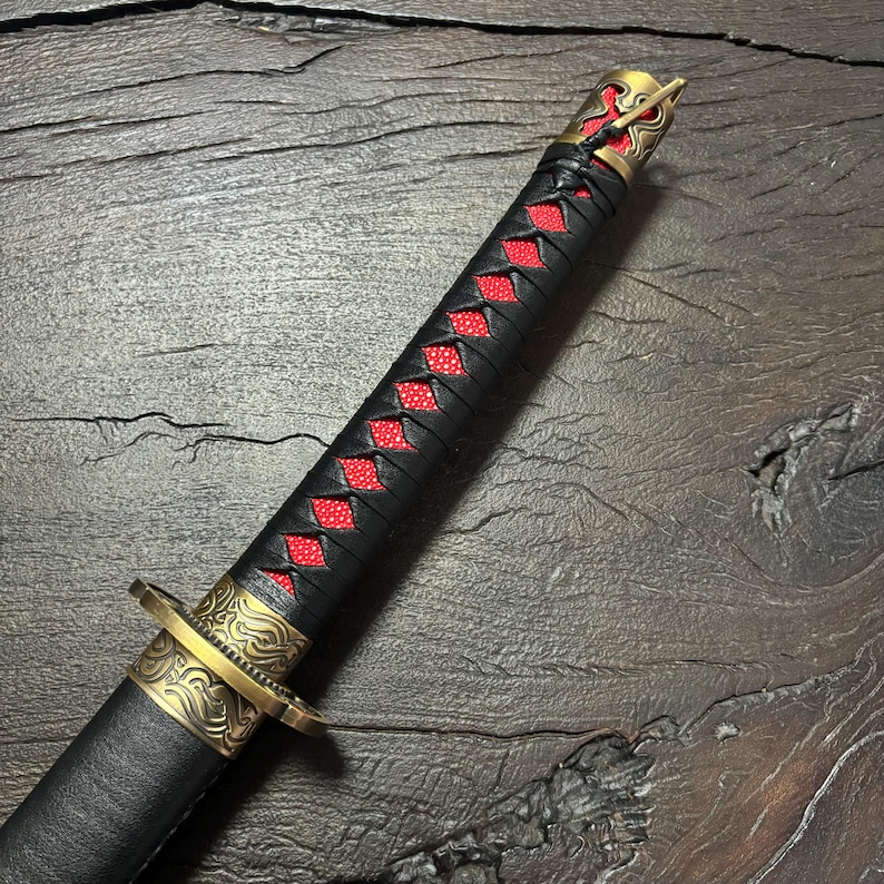 May include: Close-up of a katana sword. The handle is wrapped in black leather with red diamond-shaped accents. The guard and pommel are gold-colored with intricate designs. The blade is sheathed in a black scabbard.
