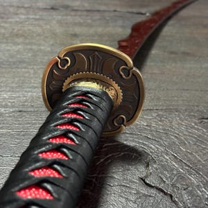May include: Close-up of a katana sword. The handle is wrapped in black fabric with red stitching. The guard is bronze-colored with intricate designs. The blade is red and curved.