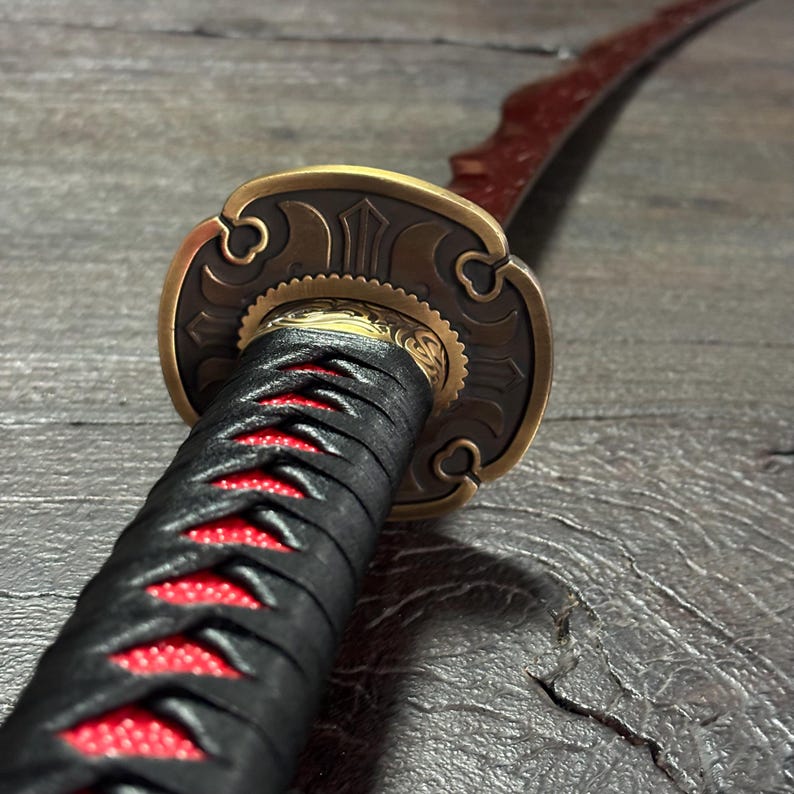May include: Close-up of a katana sword. The handle is wrapped in black fabric with red stitching. The guard is bronze-colored with intricate designs. The blade is red and curved.