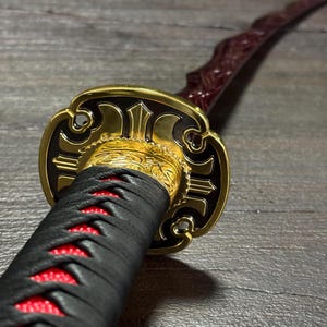 May include: A close-up of a katana sword. The hilt is wrapped in black fabric with red accents. The guard is gold and black, and the blade is a deep red color. The sword is set against a dark wood background.