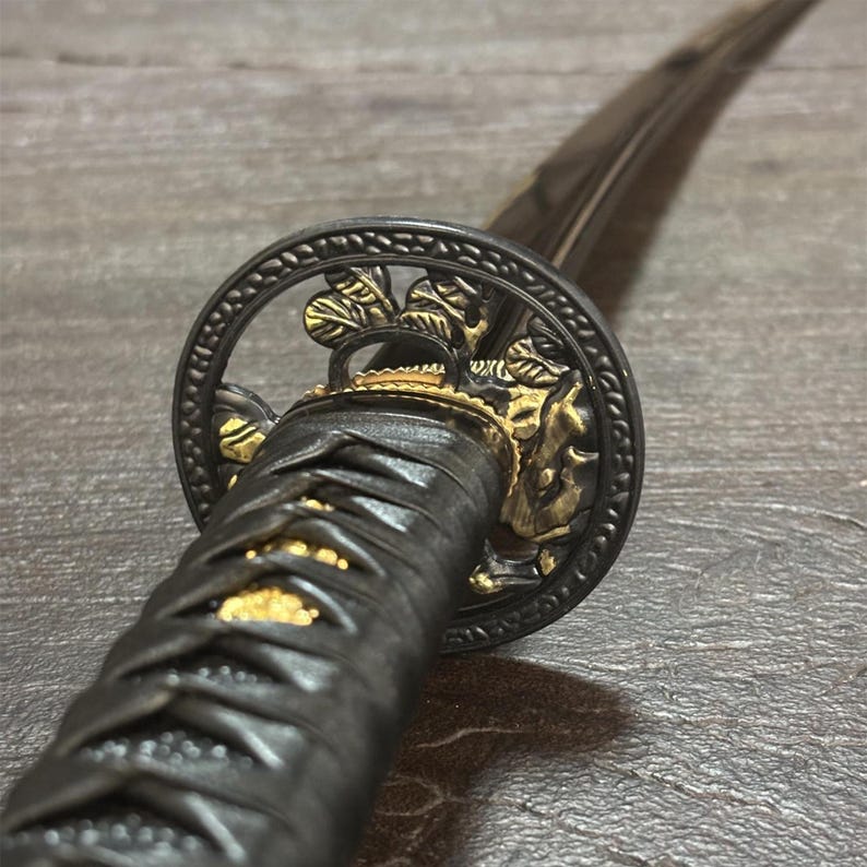 May include: Close-up of a Japanese katana sword. The hilt is wrapped in black material with gold accents. The guard is circular with a detailed floral design in gold and black. The blade is dark and slightly curved, suggesting a traditional design.