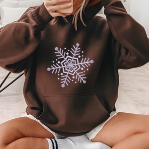 May include: A brown hoodie with a large snowflake design in white and light purple on the front. The hoodie has a kangaroo pocket and a drawstring hood. The person is wearing white shorts and white socks with navy blue stripes.