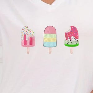 May include: White V-neck t-shirt featuring three colorful popsicle designs. The popsicles include pink with sprinkles, a pink and yellow striped popsicle, and a watermelon popsicle with a bite taken out of it.