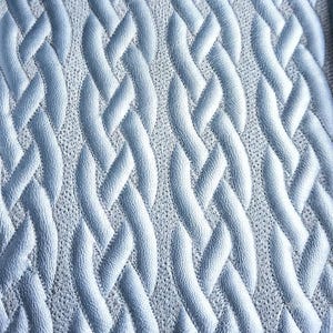 May include: A light blue fabric featuring a raised, embossed cable knit pattern. The texture consists of vertical, intertwined rope-like strands that create a three-dimensional effect against a finer, flat-knit background of the same color.