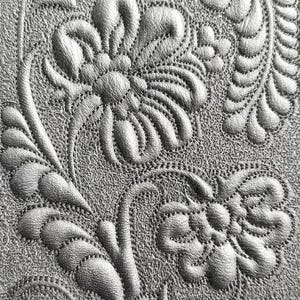May include: Close-up of a silver-colored, textured surface with an embossed floral design. The pattern features stylized flowers, leaves, and vines, with detailed stitching outlining each element. The overall effect is a detailed, tactile, and decorative pattern.