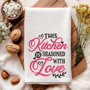 May include: White kitchen towel with black embroidery that reads "This Kitchen is Seasoned with Love" in a cursive font. The word "Love" is in pink.