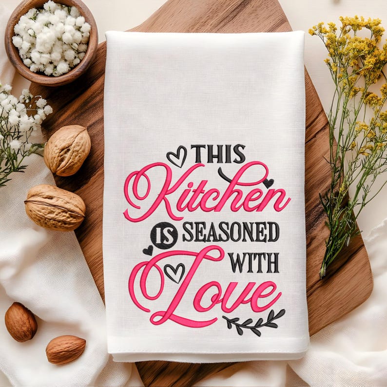 May include: White kitchen towel with black embroidery that reads "This Kitchen is Seasoned with Love" in a cursive font. The word "Love" is in pink.