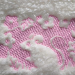 Pode incluir: Close-up de um tecido branco e macio com um design bordado em rosa. O design apresenta um rato branco rodeado por flores brancas. O fundo é branco macio e texturizado.