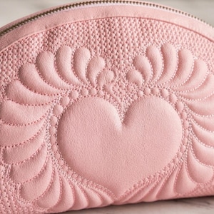 Pode incluir: Uma bolsa de cosméticos rosa claro em forma de meia-lua com um design de coração acolchoado. A bolsa apresenta uma superfície texturizada e um fecho de correr. O coração é rodeado por um padrão decorativo em forma de folha.