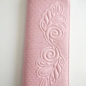 May include: A long, rectangular, light pink wallet featuring a textured, woven-like surface pattern. The front is decorated with an embossed, swirling, feather-like design created through detailed stitching that stands out against the base.