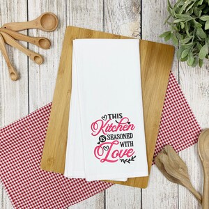 May include: White kitchen towel with the text "This Kitchen is Seasoned with Love" in black and pink lettering with a heart design.